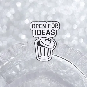 Close-up of a 'Open For Ideas' trash can enamel pin on a sparkling glass background.
