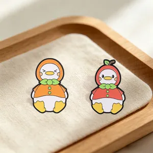 Close-up view of two cute apple and orange fruit-themed cartoon duck enamel pins on fabric