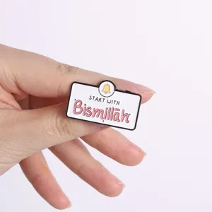 Nahaufnahme einer Emaille-Nadel mit der Aufschrift „Start With Bismillah“ und einer Glocke.