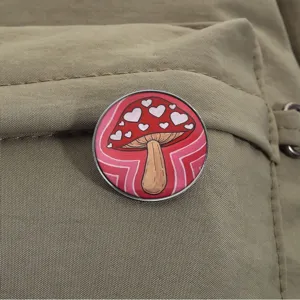 Spilla in smalto a forma di fungo rosso con cuori su sfondo di tela di zaino.