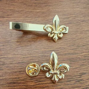 Detailed close-up of golden Fleur-de-lis tie clip and matching lapel pin on wood.