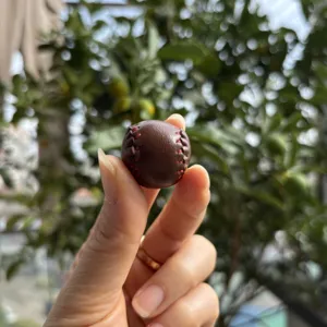 Detailed macro view of a mini brown leather baseball charm with red stitching held in hand