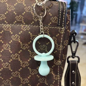 Detail eines hellblauen Schnuller-Schlüsselanhängers an einer braunen gemusterten Tasche.