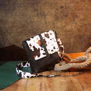 Close-up of retro Western crossbody bag with cow print flap and strap on rustic wood.