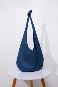 Detalle de bolso hobo denim azul con correa anudada sobre una mesa blanca.