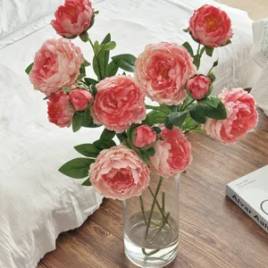Peonie artificiali rosa realistiche in un vaso di vetro su un caldo pavimento in legno.