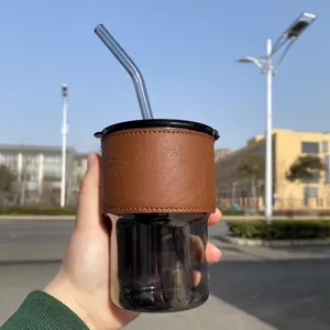 Tazza in vetro grigio fumo con custodia in pelle marrone e cannuccia tenuta in mano