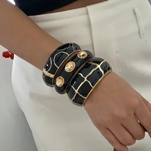 Detailansicht von drei klobigen schwarzen Emaille-Armreifen mit goldenen Mustern.