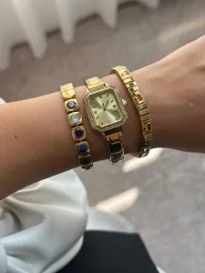 Elegante goldfarbene rechteckige Uhr mit bunten Edelstein- und Kirscharmbändern am Handgelenk.