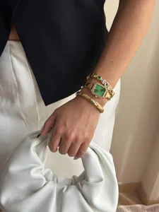 Detailaufnahme einer smaragdgrünen Golduhr und bunter Edelsteinarmbänder am Handgelenk.