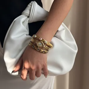 Gold evil eye watch and stacked chain bracelets worn by a model holding a white clutch bag