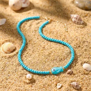 Detailansicht einer türkisfarbenen Perlenkette mit blauem Fisch-Anhänger im Sand.