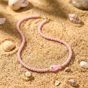 Nahaufnahme einer rosa Perlenkette im Boho-Stil mit Keramikfisch-Anhänger auf Sand.