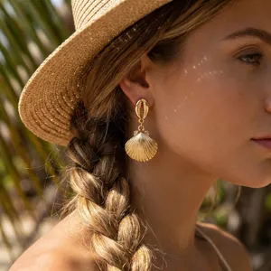 Nahaufnahme von eleganten goldenen Muschel-Ohrringen mit Strohhut für Strandurlaub-Look