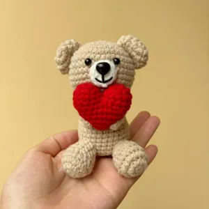 Handmade crochet teddy bear holding a red heart, detailed close-up shot of cute plush toy.