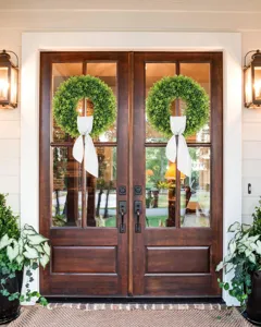 Artificial boxwood wreaths with white bows hanging on elegant wooden front doors.