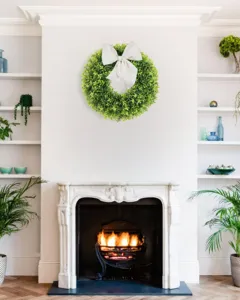 Artificial boxwood wreath with a large white bow hung above a cozy home fireplace.