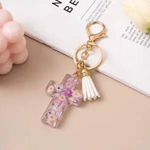 Close-up of transparent resin cross keychain with embedded purple flowers and tassel.