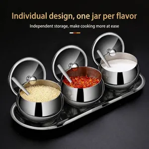 Set of 3 stainless steel seasoning jars with glass lids, spoons, and tray for kitchen storage.