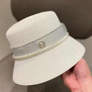 Detail view of a white straw bucket hat featuring pearl string, ribbon, and rhinestone M logo.