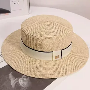Detailed view of women's beige straw boater hat with ribbon and metal M letter decor.