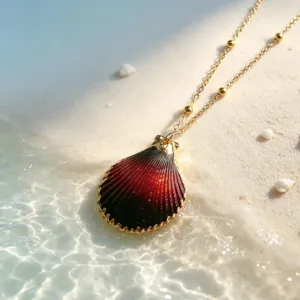 Close-up of a stunning dark red shell pendant necklace on sandy beach with water reflection.