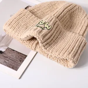 Mutstraining detail, beige gebreid, warme omslagmuts met metalen logo accent dichtbij