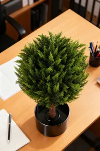 Realistic artificial cedar ball topiary in a black pot placed on a wooden office desk
