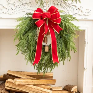 Detail of a green cedar Christmas wreath with a large red bow and bells on a white mantel.