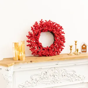 Corona navideña decorativa de bayas rojas sobre una repisa blanca con velas encendidas.