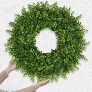 Close-up view of a lush green artificial boxwood wreath held against a white wall