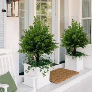 Detailed view of two artificial cedar topiary trees in white planters on a porch