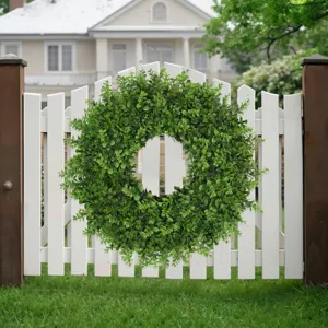 Lush green artificial boxwood wreath displayed on a white garden picket fence outdoors