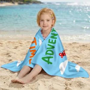 Mädchen sitzt am Strand, eingewickelt in ein blaues Mikrofaserhandtuch mit Aufdruck