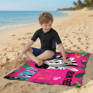 Kind sitzt auf einem pinken Mikrofaser-Strandtuch mit Zebra-Design an einem sonnigen Strand.