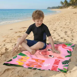 Kind sitzt auf rosa Mikrofaser-Strandtuch mit Schweinchen-Design am sonnigen Strand.