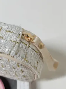 Détail rapproché d'une trousse à maquillage en tweed blanc avec fermeture éclair dorée.