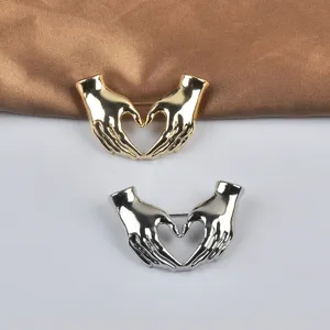 Detalle de broches de metal en oro y plata con forma de gesto de corazón con ambas manos