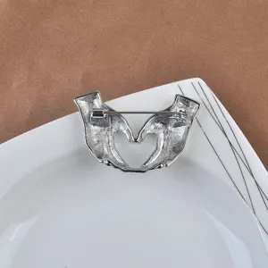 Vista detallada del revés de un broche plateado con forma de manos en corazón y alfiler.