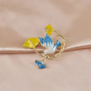 Elegante Brosche mit Emaille-Friedenstaube und Ginkgoblatt auf rosa Satinhintergrund.