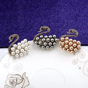 Three elegant swan brooches in white, grey, and champagne pearls on a white plate