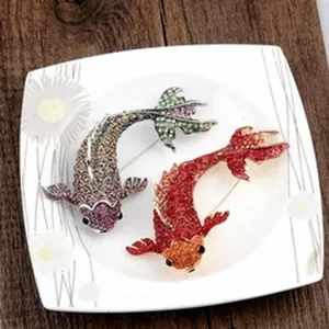 Detail showing two rhinestone-encrusted koi fish brooches in red and silver on a white plate.