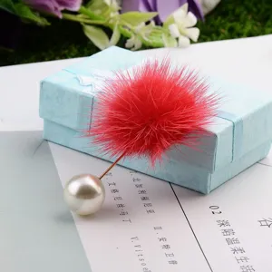 Detailansicht einer roten Nerzfell-Pompon-Brosche mit Perle auf einer blauen Box