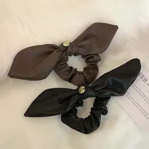 Eleganti elastici per capelli in ecopelle con fiocco e ciondolo oro su sfondo morbido.