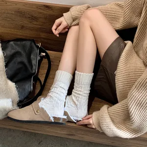 Close-up of white cable-knit cashmere socks worn with tan mules on a wooden bench.