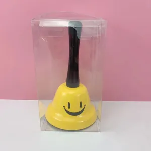 Campana de mano amarilla con cara sonriente negra y mango en caja transparente para juegos.