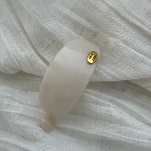 Eleganter cremeweißer ovaler Haarclip mit goldenem Hand Made Tag auf Stoffstruktur