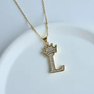 Collier lettre L couronne dorée en gros plan, pendentif initial en cristal sur plateau blanc.
