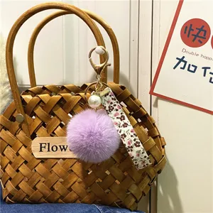 Detail of purple fur pom-pom keychain with floral strap and pearl accent on woven bag.