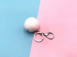 Close-up of a fluffy white pom pom keychain with a silver metal clasp on a pink and blue background.